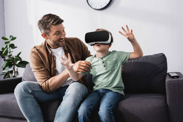 smiling father touching surprised son using vr headset and gesturing whiles sitting on sofa - Foto, afbeelding