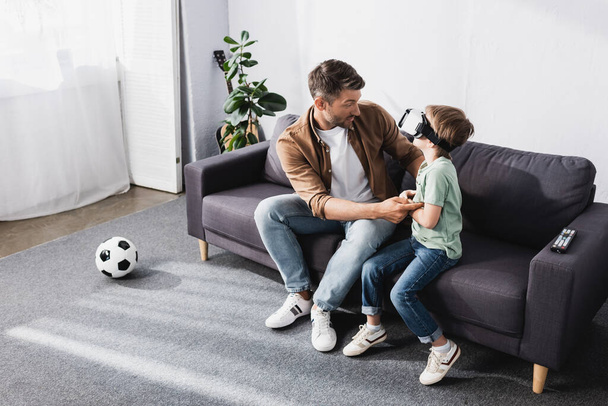 aus der Vogelperspektive: Mann berührt Sohn mit Kopfhörer, während er auf Sofa sitzt - Foto, Bild
