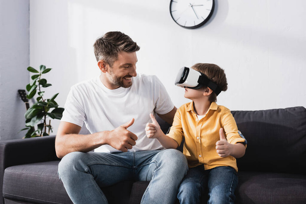 smiling man and son in vr headset showing thumbs up while sitting on sofa - Фото, изображение