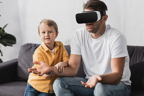 cute boy touching hand of father in vr headset showing shrug gesture - Foto, imagen