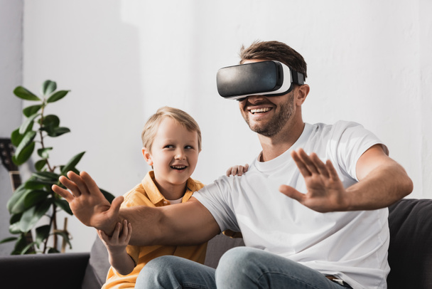 smiling father in vr headset gesturing while cute son sitting on sofa - Foto, Bild