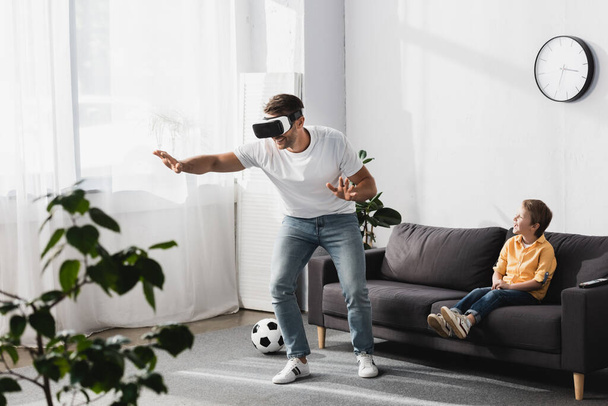 selective focus of man in vr headset walking and gesturing while adorable son sitting on sofa - Foto, afbeelding