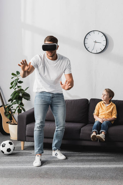 man in vr headset walking abd gesturing while adorable son sitting on sofa - Photo, image