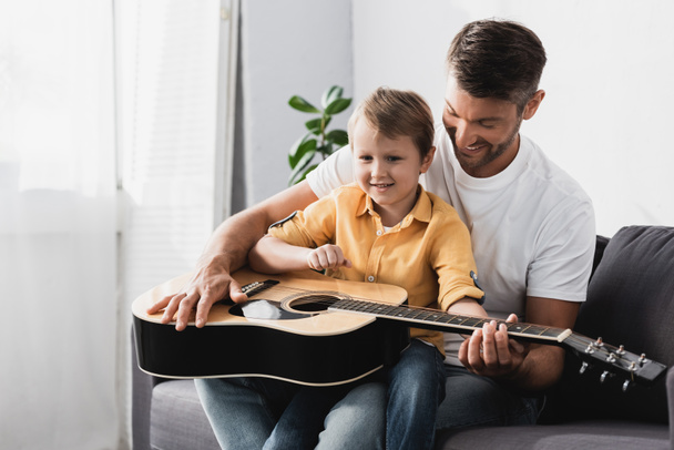smiling boy sitting of fathers knees and learning how to play acoustic guitar - Photo, Image
