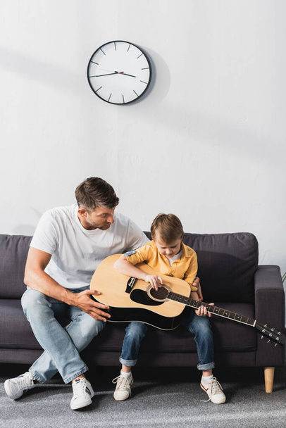 uważny chłopiec uczy się grać na gitarze akustycznej siedząc na kanapie obok ojca - Zdjęcie, obraz