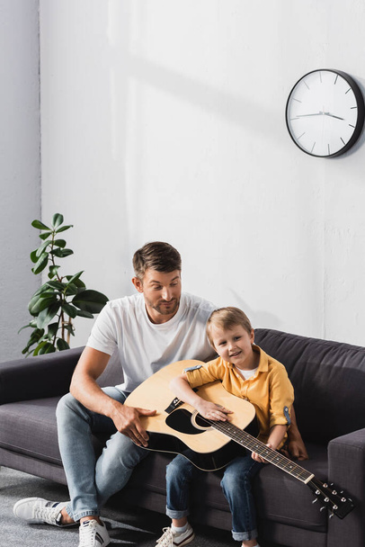 όμορφος πατέρας μαθαίνει στο γιο του να παίζει ακουστική κιθάρα στον καναπέ στο σπίτι - Φωτογραφία, εικόνα