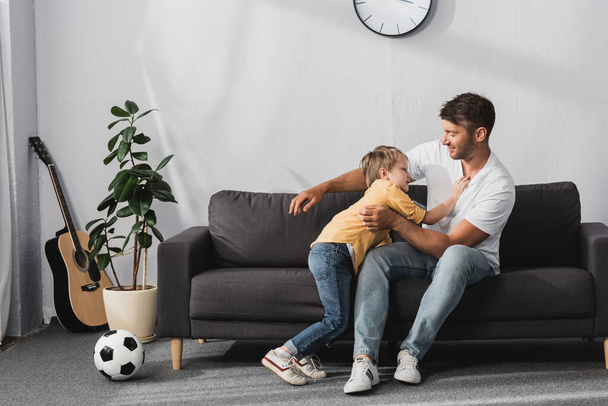 padre y adorable hijo en broma luchando en el sofá cerca de pelota de fútbol y maceta planta
 - Foto, Imagen