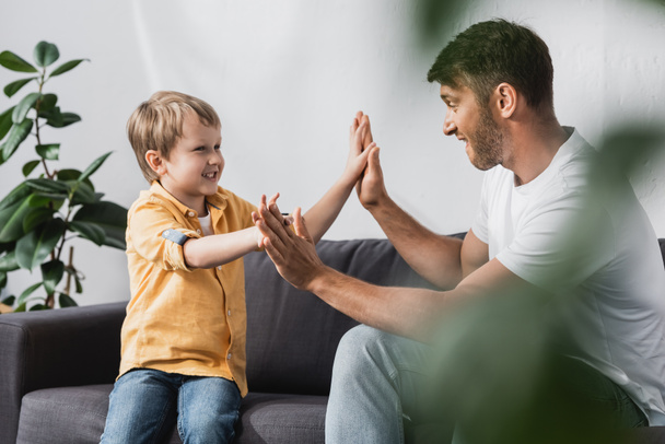 selectieve focus van vrolijke vader en zoon geven high five tijdens het zitten op de bank thuis - Foto, afbeelding