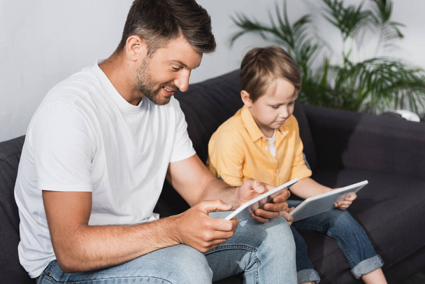 selective focus of smiling father and attentive son using digital tablets at home - Фото, изображение