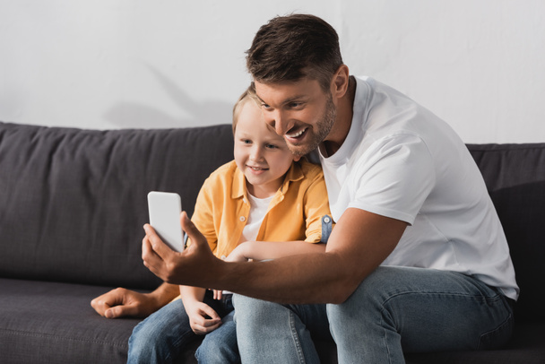 smiling father and son taking selfie on smartphone while sitting on sofa - Foto, Imagen