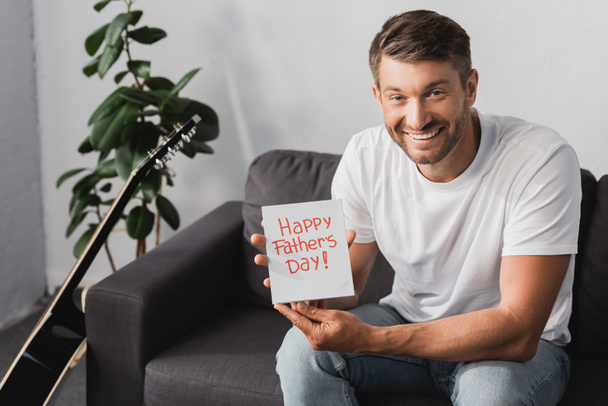 homem feliz mostrando feliz pai cartão de dia enquanto sentado no sofá perto de planta e guitarra acústica
 - Foto, Imagem