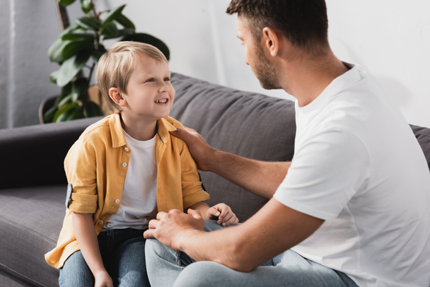 father touching shoulder of smiling son while talking to him on sofa - Φωτογραφία, εικόνα