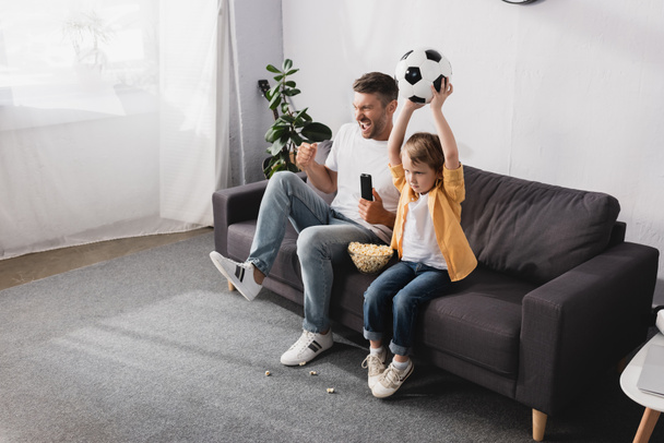 excited man showing winner gesture near son holding soccer ball in raised hands while watching tv - Foto, Imagen