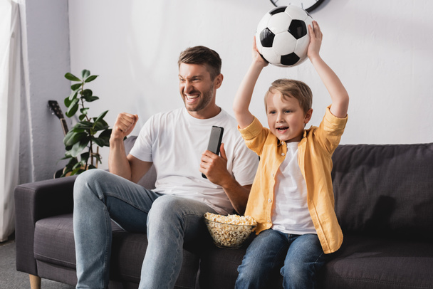 excited man showing winner gesture near son holding soccer ball in raised hands while watching tv - Foto, immagini