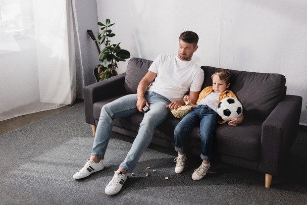 βαριεστημένος πατέρας και γιος κάθονται στον καναπέ με ποπ κορν και μπάλα ποδοσφαίρου και βλέποντας τηλεόραση - Φωτογραφία, εικόνα
