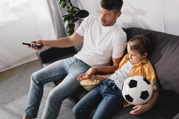 Yüksek açılı, elinde futbol topu olan bir çocuğun TV izlerken babasıyla patlamış mısır yediği görüntüsü. - Fotoğraf, Görsel