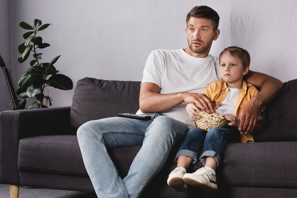 attentive father and son watching tv and eating popcorn on sofa at home - Photo, Image