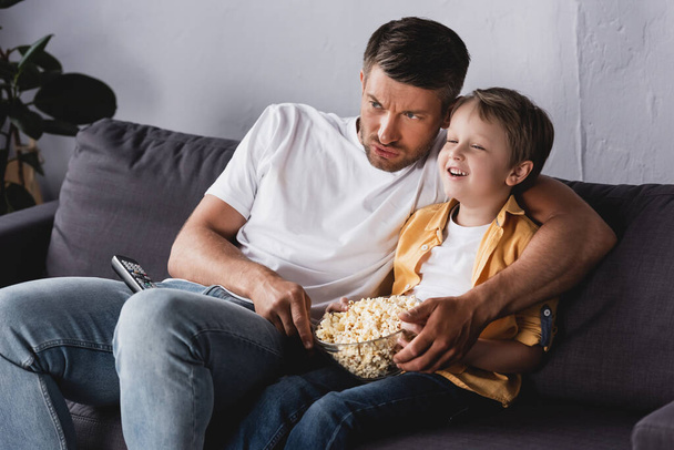 père inquiet et fils souriant regarder la télévision et manger du pop-corn sur le canapé à la maison - Photo, image