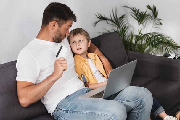 apa kezében hitelkártya és nézi aranyos fiú, miközben ül a kanapén laptop - Fotó, kép