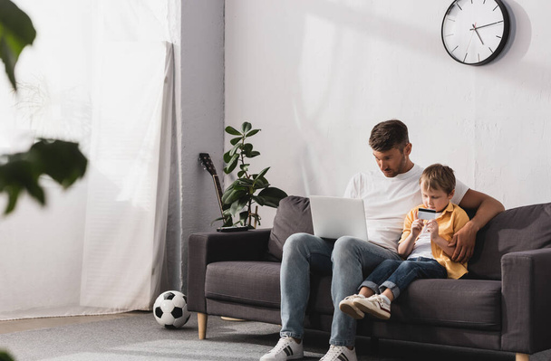 enfoque selectivo de padre usando el ordenador portátil y su hijo con tarjeta de crédito mientras está sentado en el sofá en casa
 - Foto, Imagen