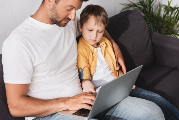 attentive father and son using laptop while sitting on sofa at home - Photo, Image