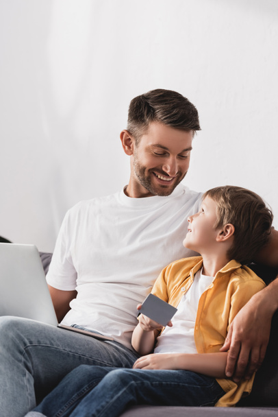 щасливий батько з ноутбуком дивиться на усміхненого сина, який тримає кредитну картку
 - Фото, зображення
