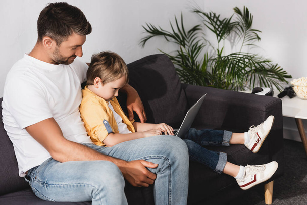 adorable chico usando laptop mientras sentado en sofá junto con padre
 - Foto, Imagen