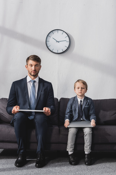 serieuze zakenman en zijn zoon in formele kleding zitten met laptops op de bank thuis en kijken naar de camera - Foto, afbeelding