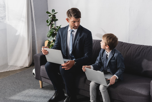 serious businessman with his son in formal wear sitting on sofa with laptops - Foto, Imagen