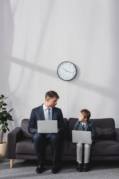 серйозний бізнесмен і його син в офіційному одязі сидить на дивані з ноутбуками і дивиться один на одного
 - Фото, зображення