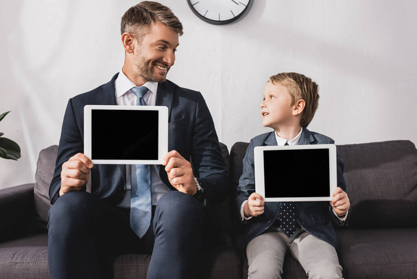 χαμογελαστός επιχειρηματίας και γιος με επίσημη ένδυση που δείχνει ψηφιακές ταμπλέτες με λευκή οθόνη ενώ κοιτάζονται μεταξύ τους - Φωτογραφία, εικόνα
