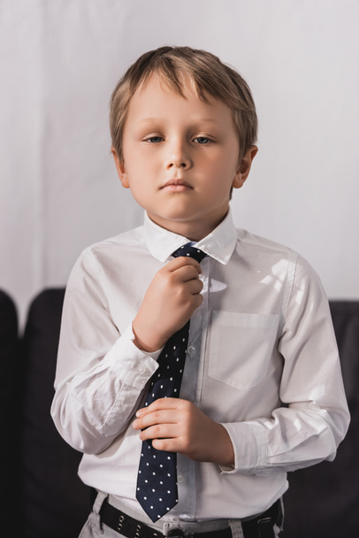 serious, cute boy in white shirt putting tie on and looking at camera - Fotografie, Obrázek