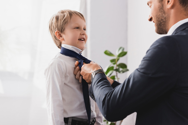 businessman in formal wear putting tie on happy son at home - 写真・画像
