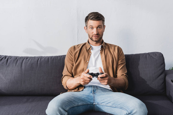 KYIV, UKRAINE - JUNE 9, 2020: concentrated man sitting on sofa and playing video game with joystick - Photo, image