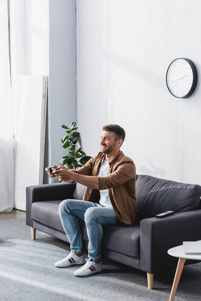 KYIV, UKRAINE - JUNE 9, 2020: excited man playing video game while sitting on sofa - Foto, afbeelding