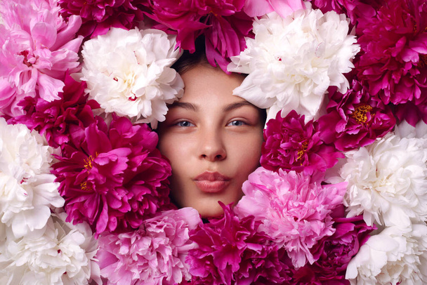 Attractive female with rounded lips looking at camera, lady with beautiful eyes, face with pink, white and burgundy peony flowers, charming woman showing kiss gesture. - Photo, Image