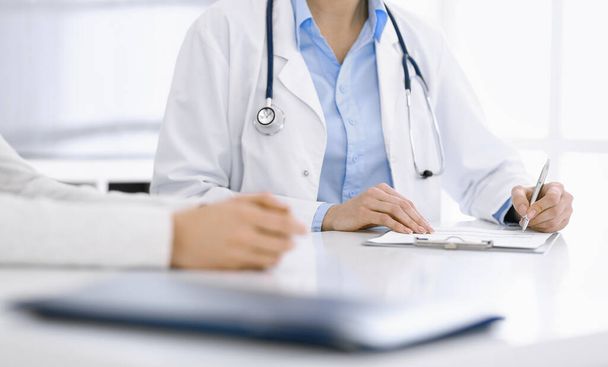 Unknown woman-doctor and female patient sitting and talking at medical examination in clinic, close-up. Therapist wearing blue blouse is filling up medication history record. Medicine concept - Photo, Image