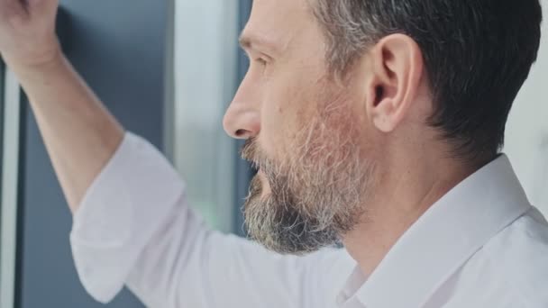 A close-up view of a serious mature man is looking outside while standing near the window in the office - Filmmaterial, Video