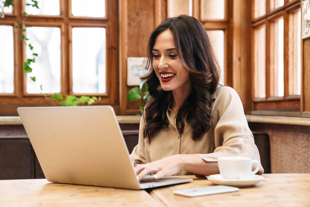 Image of happy brunette adult woman smiling and working with laptop while sitting in cozy cafe indoors - 写真・画像