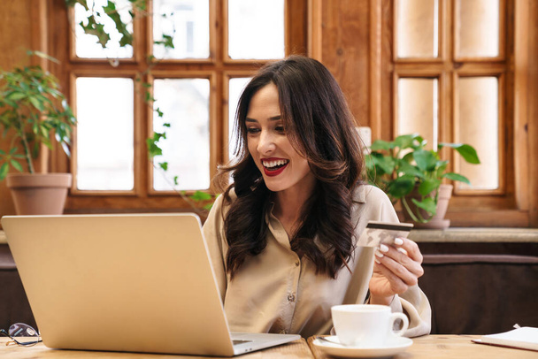 Image of excited brunette adult woman holding credit card and working with laptop while sitting in cafe indoors - Foto, imagen
