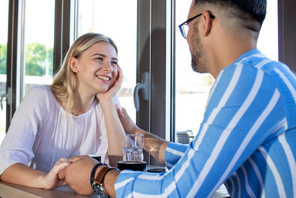 Молодая романтическая пара, наслаждающаяся чашечкой кофе в кафе, сидя за столом, улыбаясь и разговаривая. Парень нежно трогает свою девушку.
. - Фото, изображение
