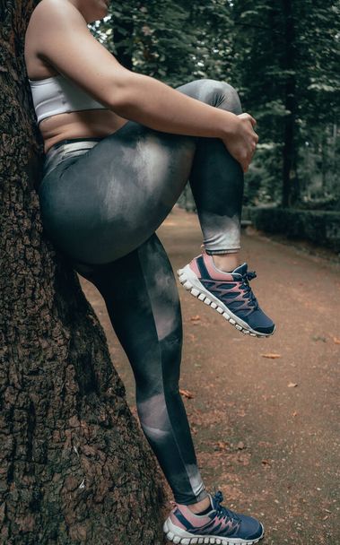 Athlète méconnaissable étirant les jambes sur le terrain. Récolte d'athlète élégant étirant les jambes dans une forêt appuyée contre un arbre. - Photo, image