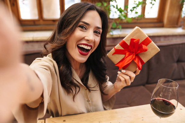 Image of excited adult woman taking selfie photo with gift box while sitting in cozy cafe indoors - Foto, Imagem