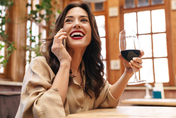 Image of cheerful adult woman drinking red wine and talking on mobile phone while sitting in cafe indoors - Photo, image