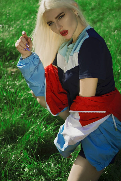 Молодая красивая девушка с светлыми волосами в разноцветной спортивной форме на фоне зеленой травы
 - Фото, изображение