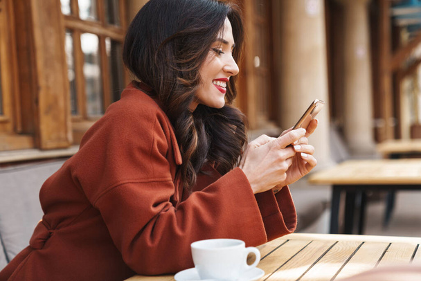 Image of happy beautiful adult woman using cellphone and drinking coffee while sitting in street cafe outdoors - 写真・画像
