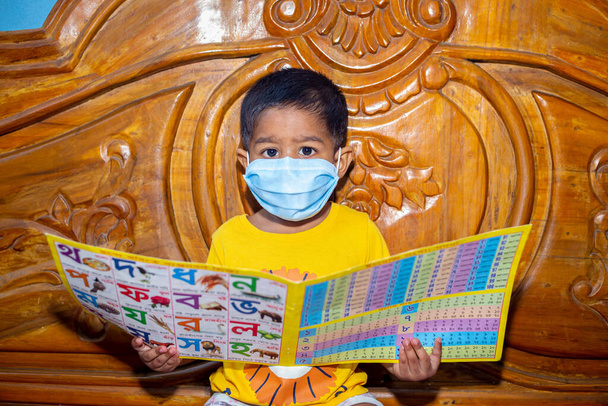 Ein kleiner Junge trägt eine Gesichtsmaske und sitzt auf einem Bett und liest ein Buch. Gesichtshygienische Maske für Virensicherheit. Babyporträt in häuslicher Quarantäne. - Foto, Bild
