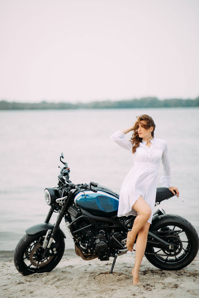 Mladá žena stojí na pláži v bílých šatech v blízkosti motocyklu proti vodě pozadí. - Fotografie, Obrázek