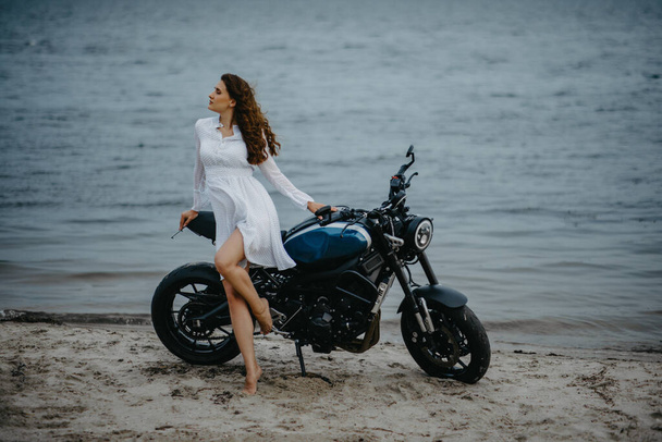 Νεαρή γυναίκα στέκεται στην παραλία με λευκό φόρεμα κοντά σε μοτοσικλέτα με φόντο το νερό. - Φωτογραφία, εικόνα
