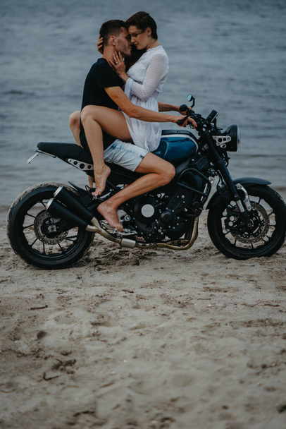 Couple in love hugs on beach sitting on motorcycle on water background. - Photo, Image